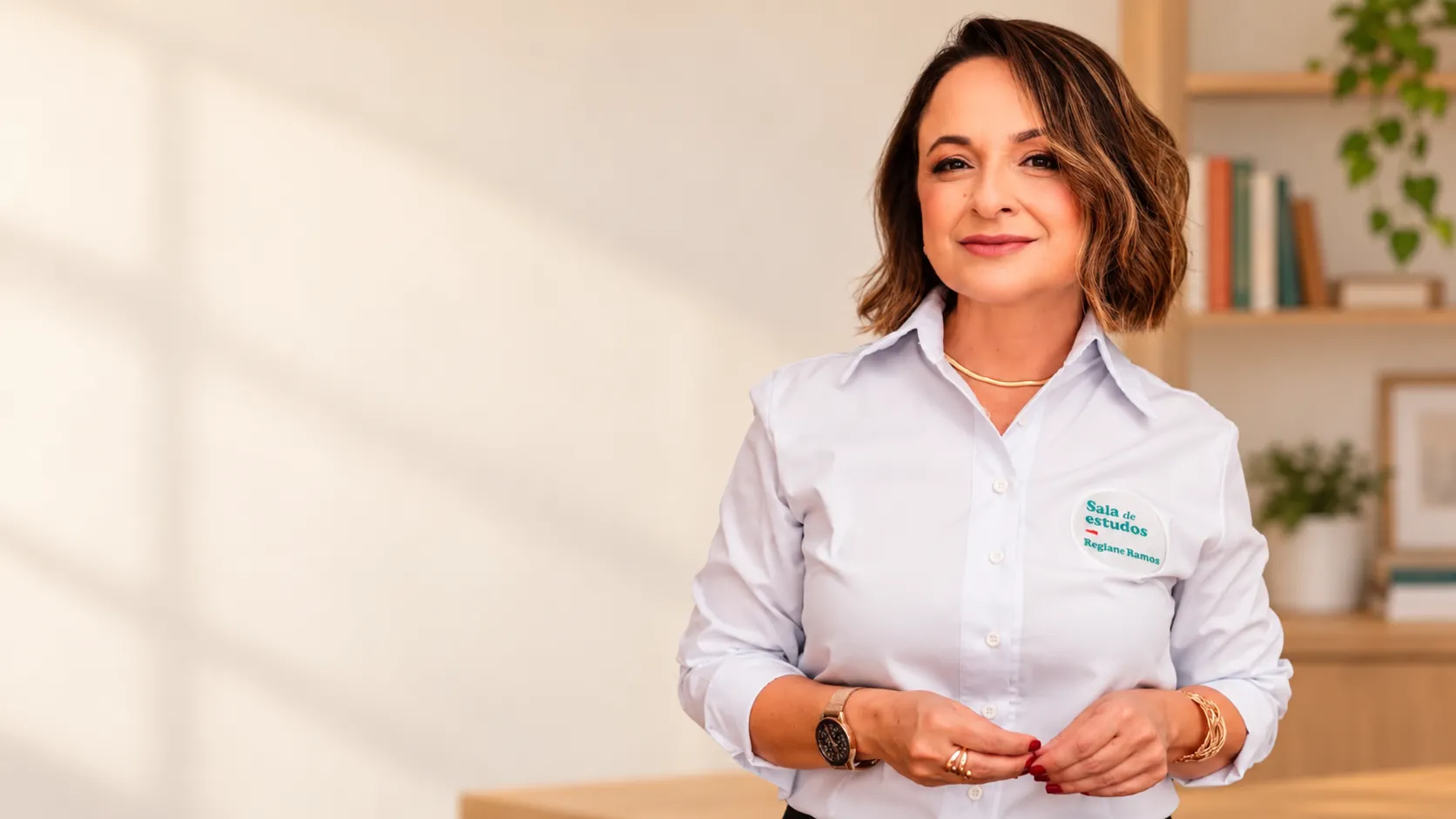Prof. Regiane Ramos sorrindo, de camisa da Sala de Estudos, em um ambiente claro com estante de livros