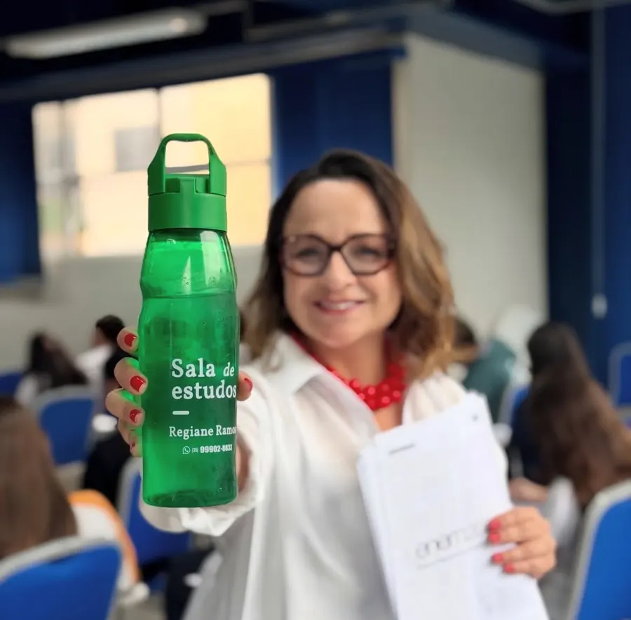Prof. Regiane em sala, mostrando a garrafa da Sala de Estudos com alunos ao fundo